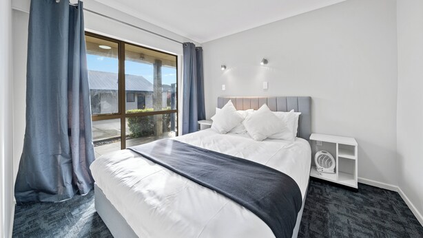 A modern hotel room with a large bed, a bedside table with a fan, and a view through a sliding glass door.