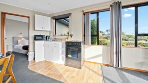 A compact kitchen with white cabinets, a microwave, a stove, and a sink. There is a dining area with a wooden table and chairs. The room has large windows with curtains, offering a view of greenery outside.