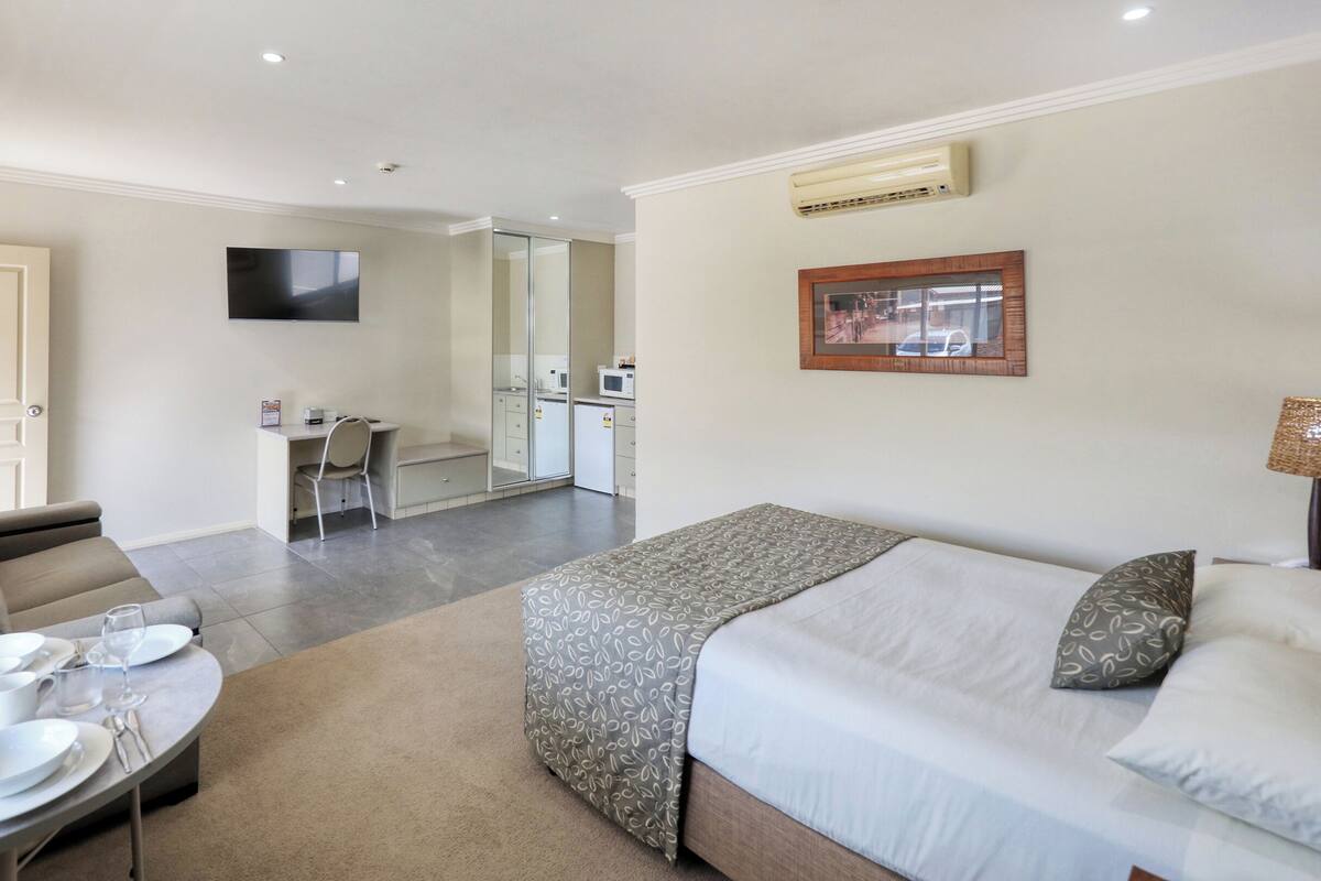 A hotel room with a bed, a sofa, a dining table set with plates and glasses, a kitchen area, and a TV mounted on the wall.