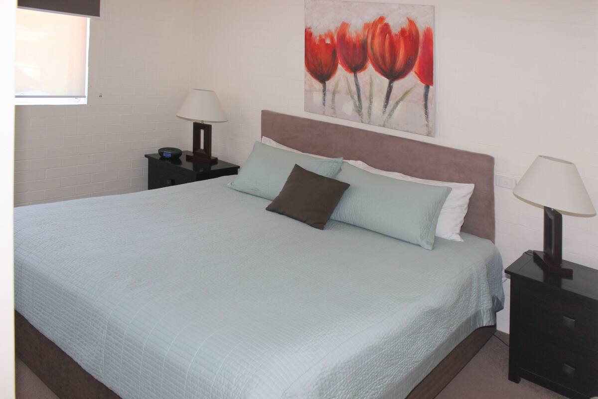 A neatly made bed with a light blue bedspread and two pillows, a nightstand with a lamp, and a painting of red tulips on the wall.