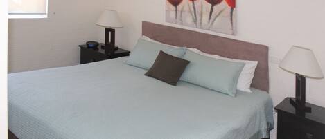 A neatly made bed with a light blue bedspread and two pillows, a nightstand with a lamp, and a painting of red tulips on the wall.