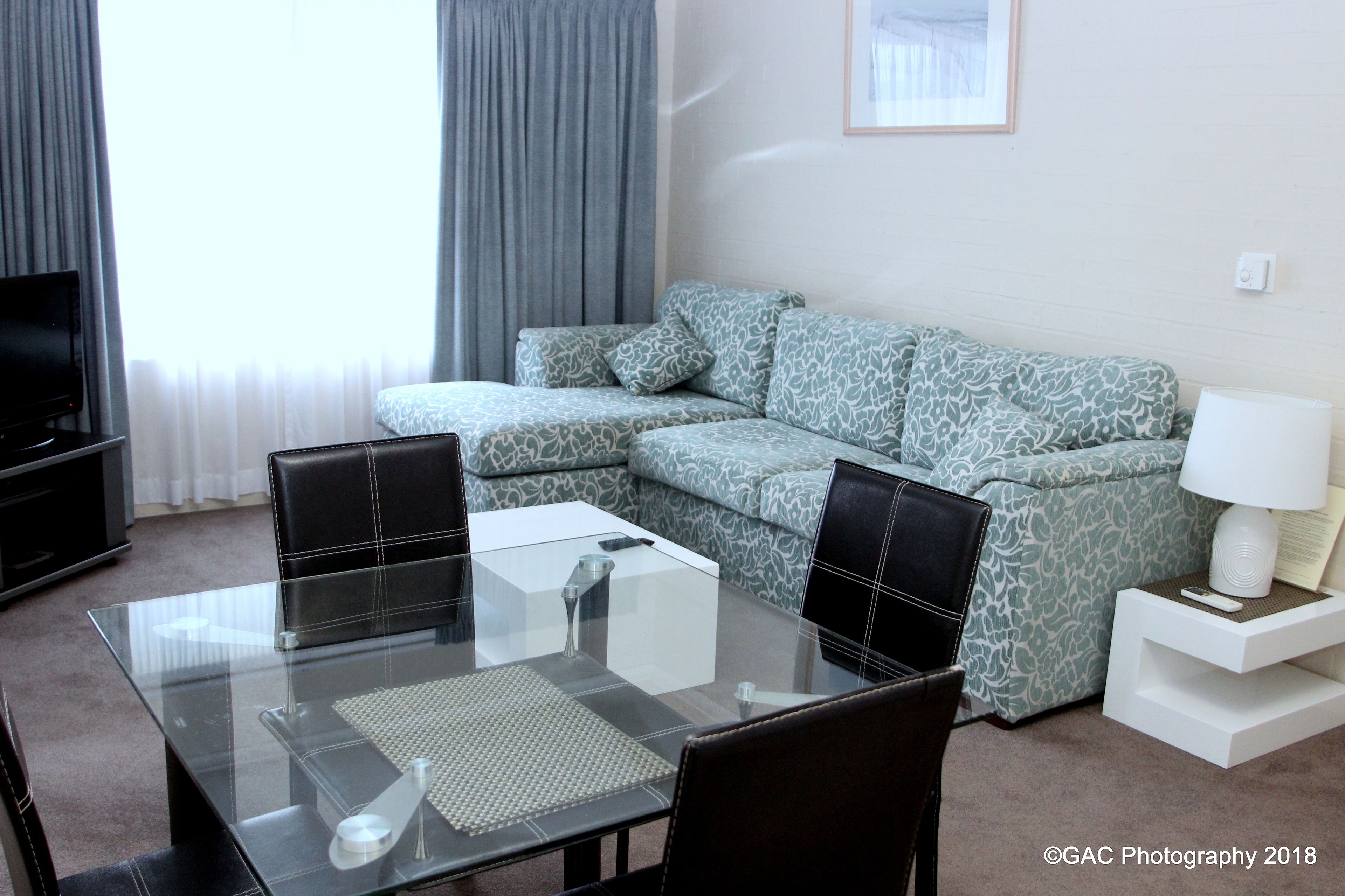 A modern living room with a glass dining table, black chairs, a patterned sofa, a TV, and a lamp on a side table.