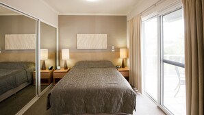 A hotel room with a large bed, a bedside table with a lamp, a nightstand, a mirror, and a window with curtains. - Mollymook Cove Apartments (Mollymook)