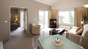 A spacious living area with a sofa, a coffee table, and a television. There is a door leading to a bedroom. - Mollymook Cove Apartments (Mollymook)