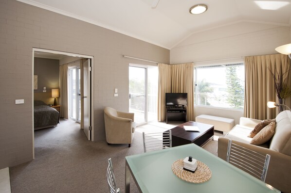 A spacious living area with a sofa, a coffee table, and a television. There is a door leading to a bedroom. - Mollymook Cove Apartments (Mollymook)