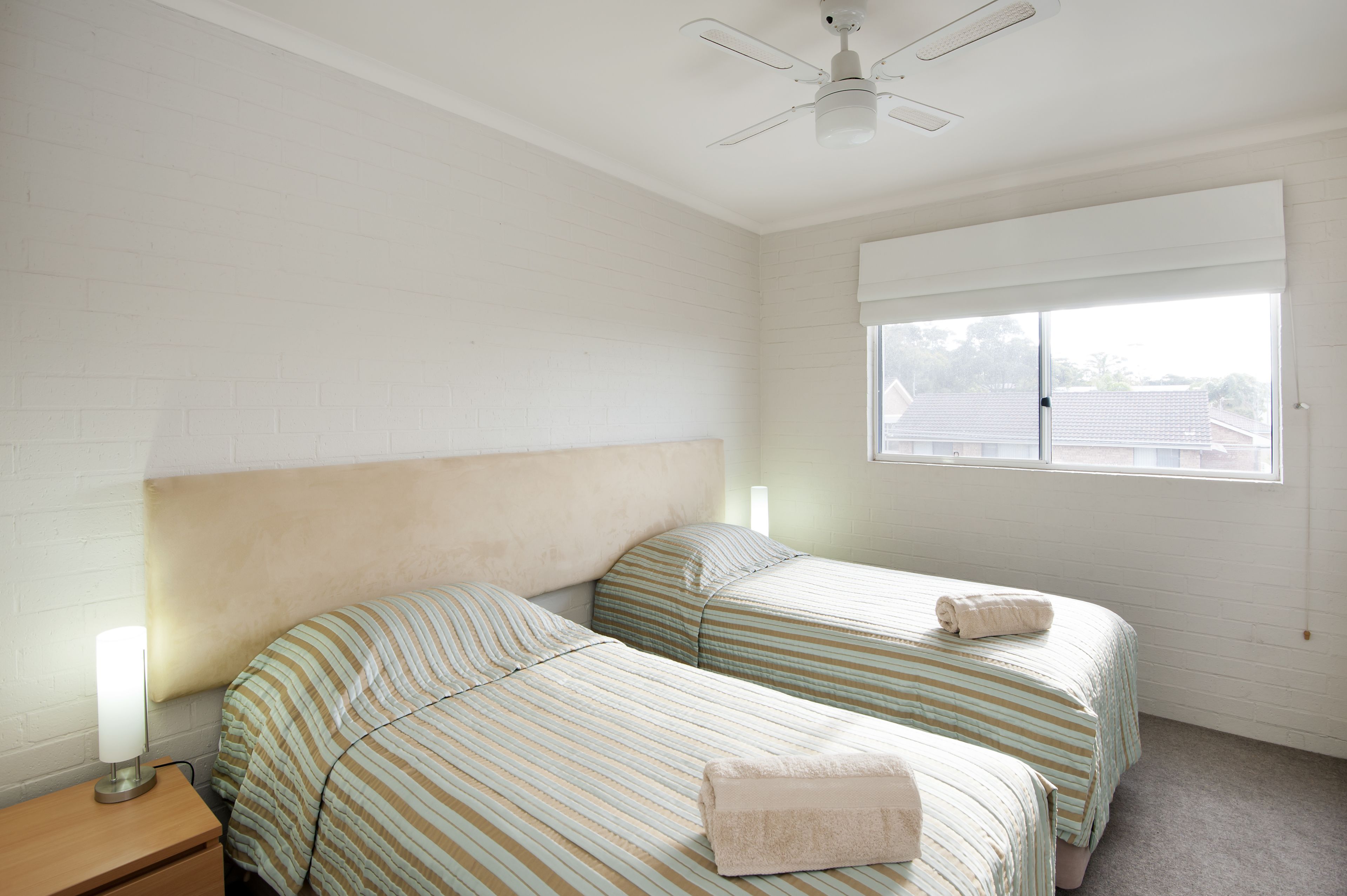 A room with two beds, a ceiling fan, a window with blinds, and a bedside lamp.