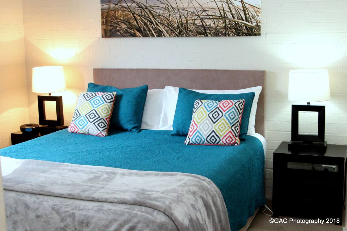 A neatly made bed with a teal bedspread and patterned pillows, flanked by two bedside tables with lamps, in a room with a white brick wall and a decorative plant.
