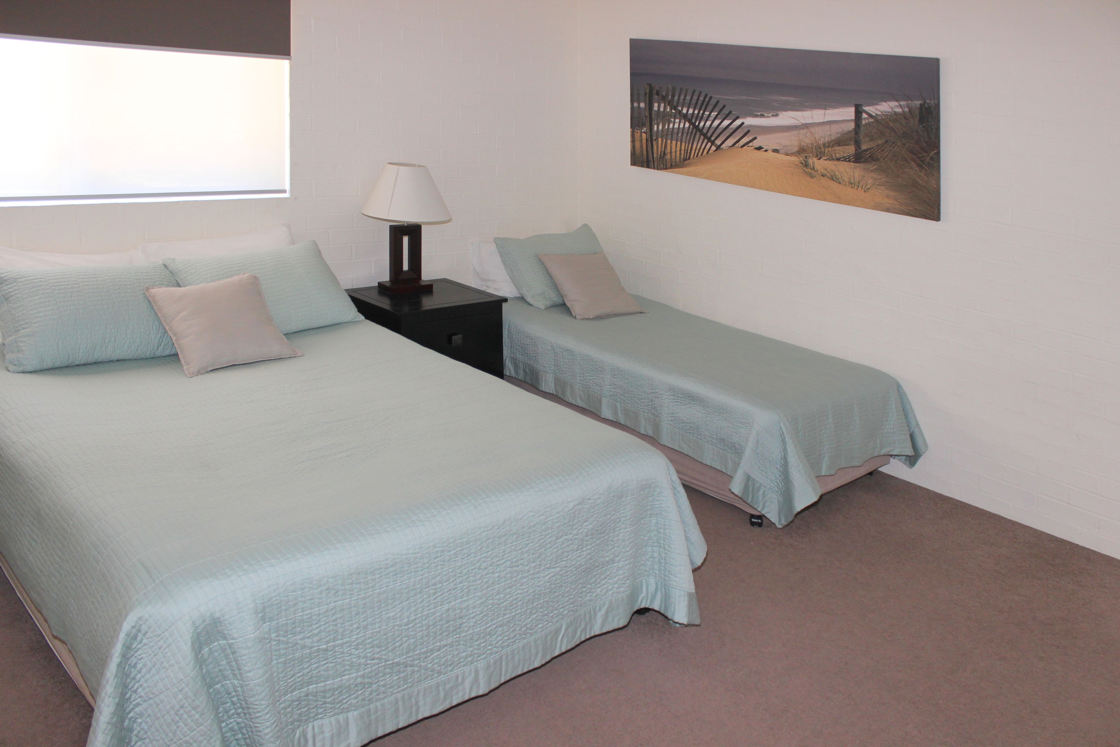 A room with two beds, each with a light blue bedspread and pillows, a nightstand with a lamp, and a beach-themed picture on the wall.