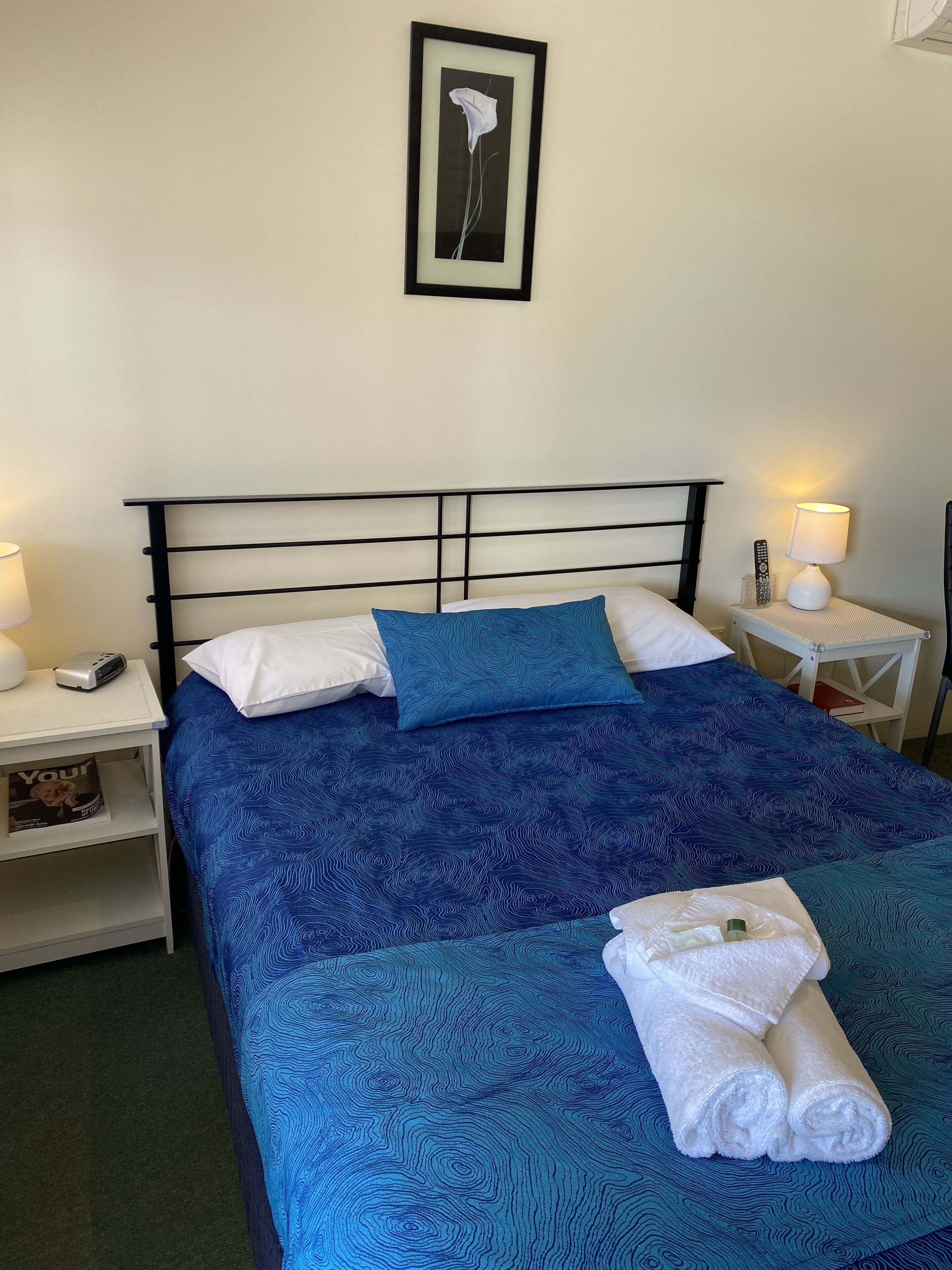 A neatly made bed with a blue patterned bedspread, white pillows, and a framed artwork above it.