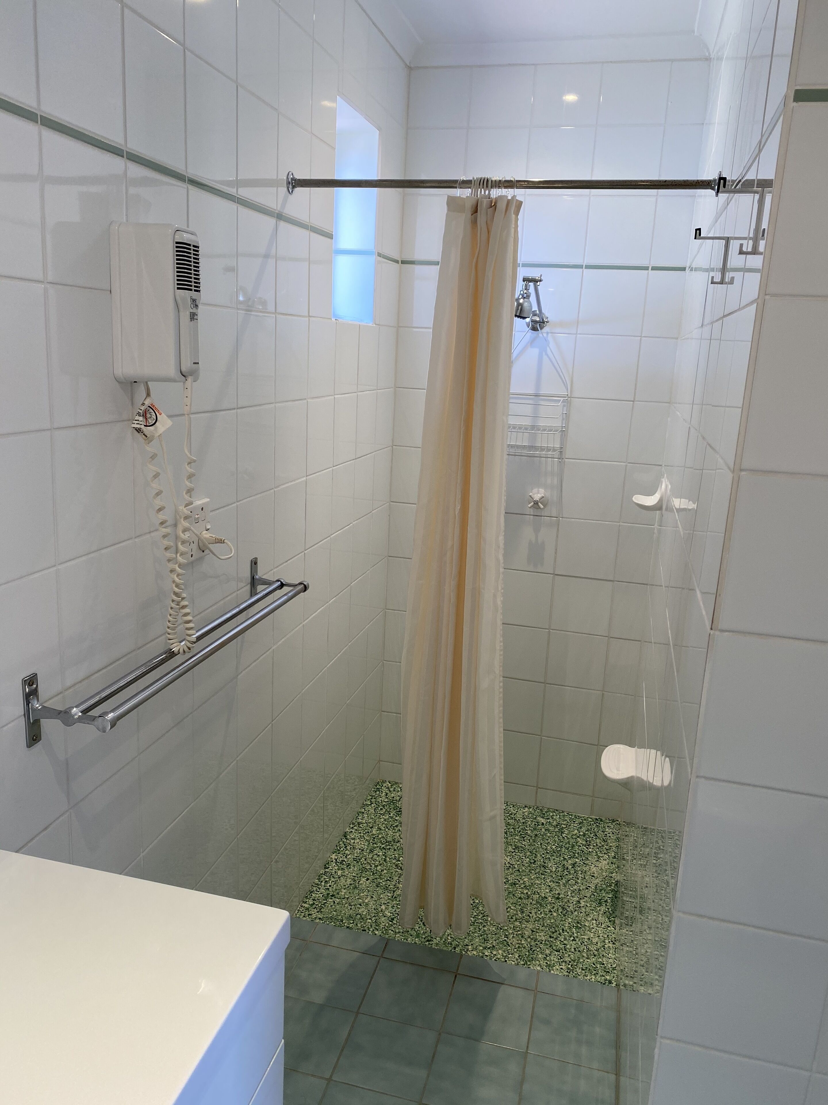 A modern bathroom with white tiles, a glass shower enclosure, a beige shower curtain, a handheld showerhead, and a towel rack.