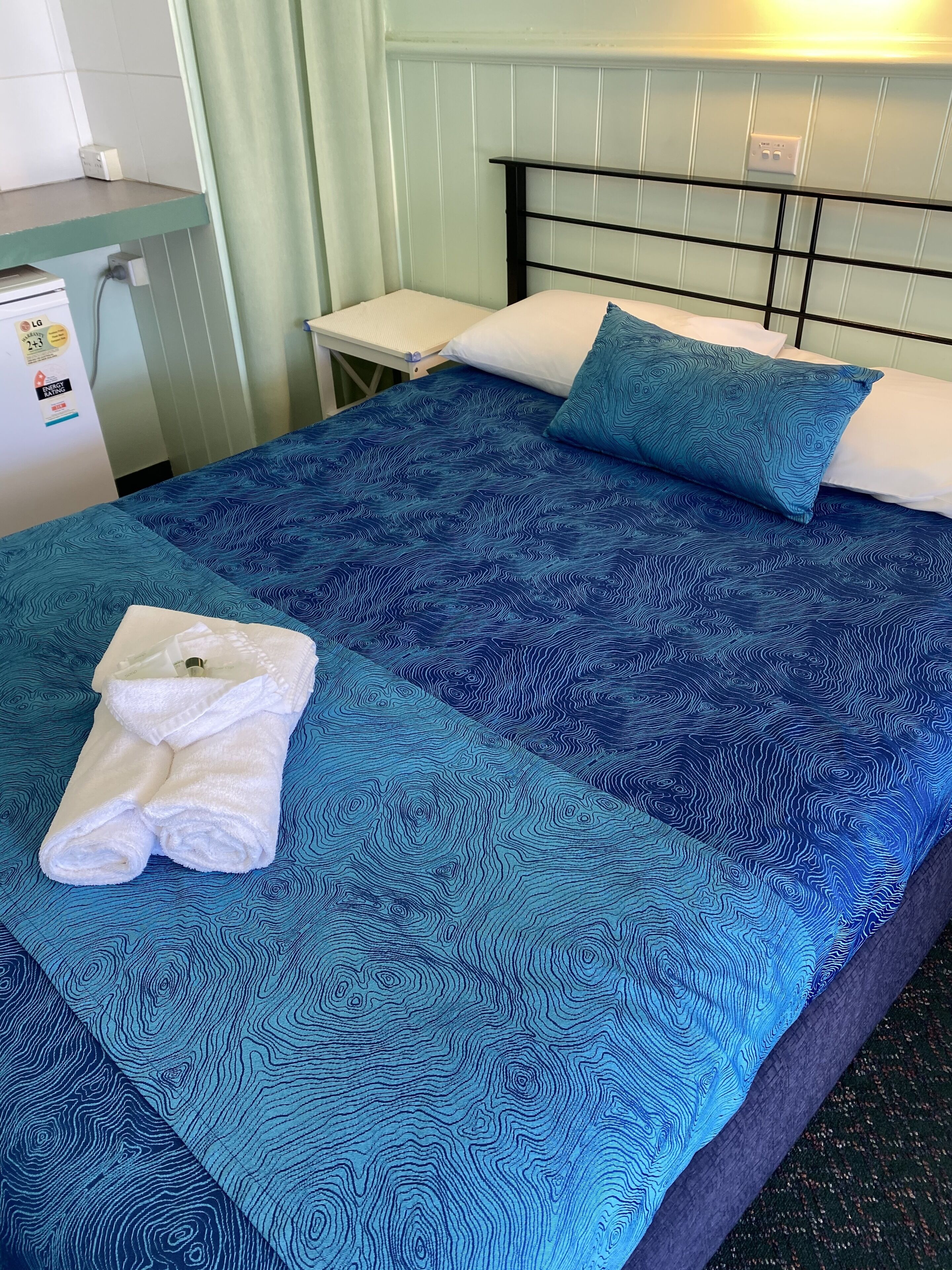 A bed with a blue patterned bedspread, a white pillow, a blue pillow, and a folded white towel on the bed.