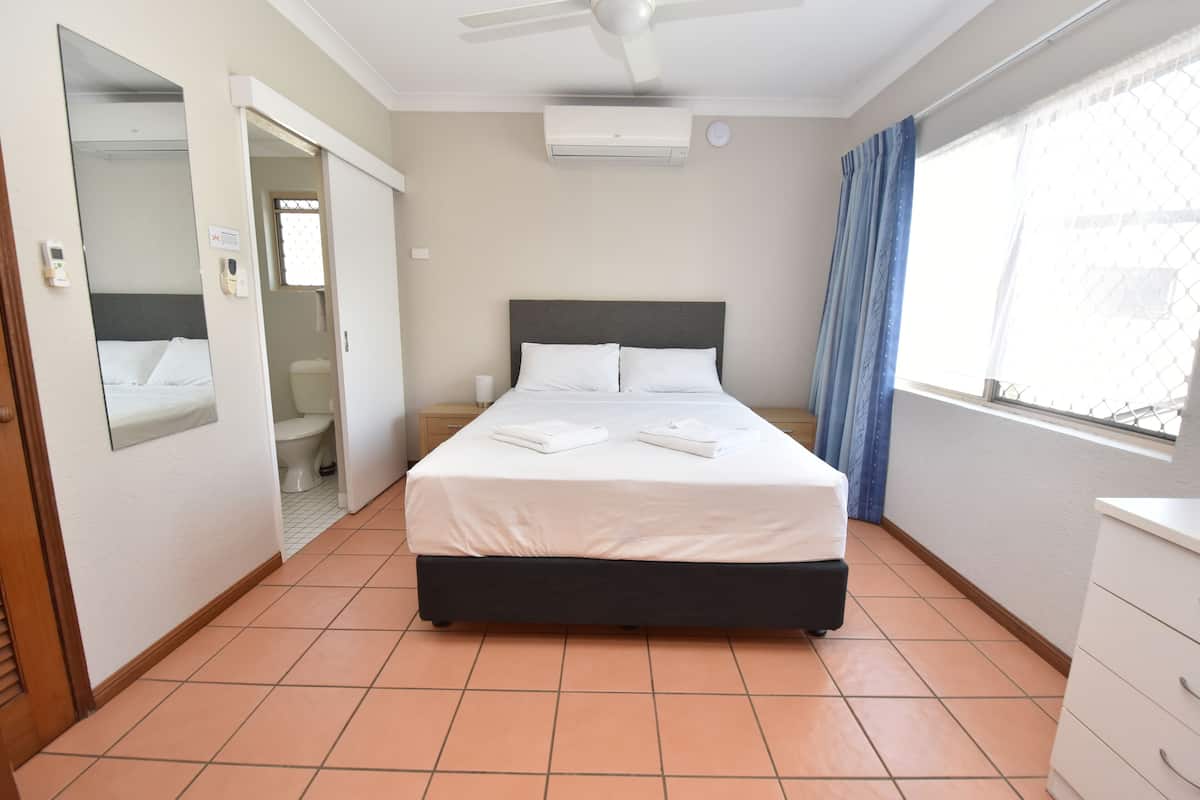 A hotel room with a bed, a ceiling fan, an air conditioning unit, a window with curtains, and a bedside table.