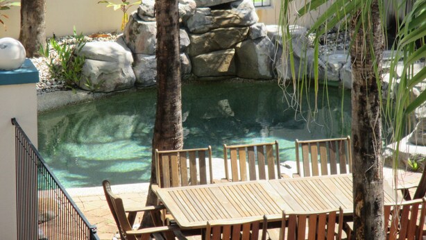 Zona de piscina con mesa y sillas de madera, rodeada de rocas y palmeras.