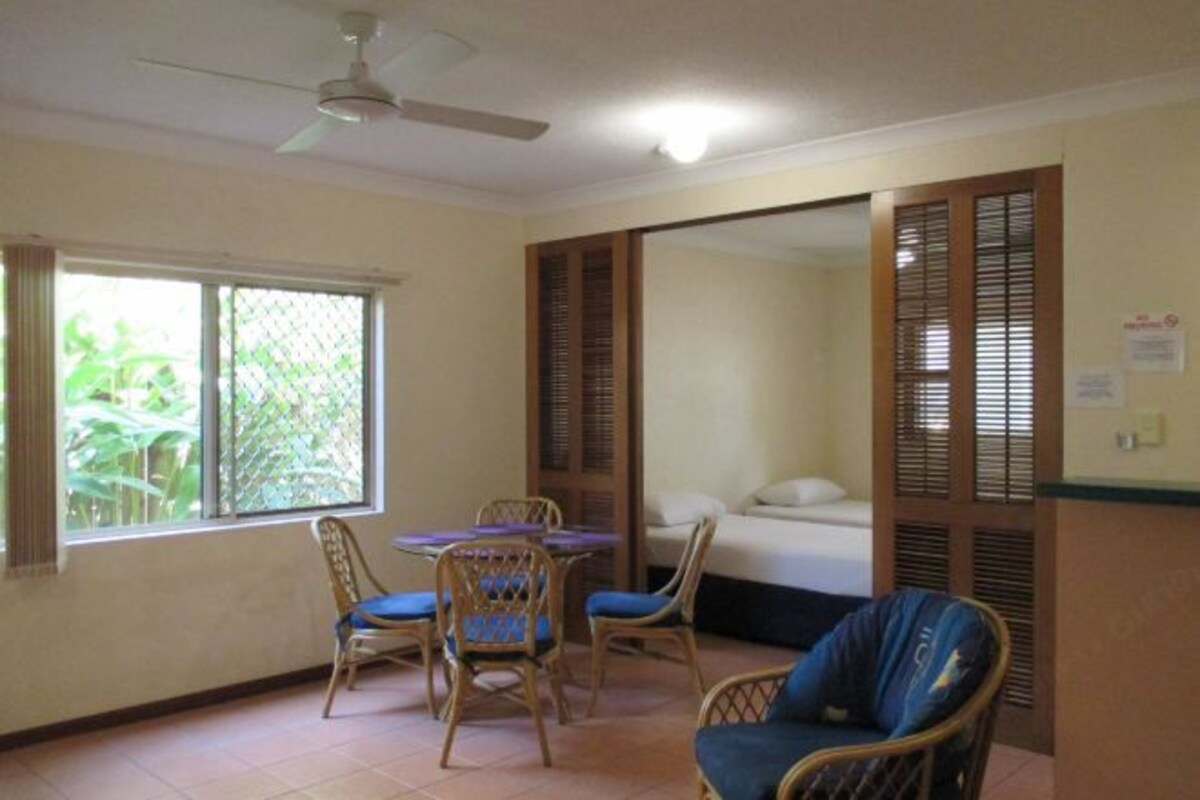 Una habitación con cama, una mesa de comedor con sillas, un ventilador de techo y una ventana con persianas.