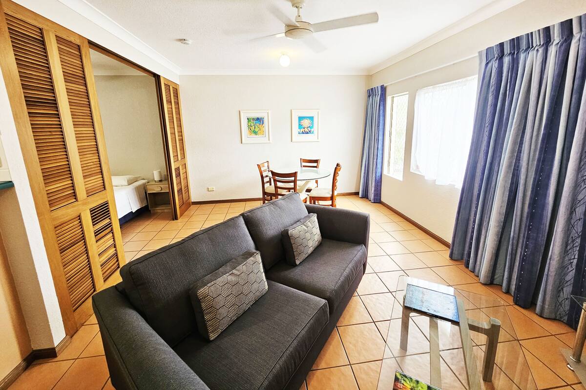 A living room with a sofa, a ceiling fan, a dining table with chairs, and a window with curtains.