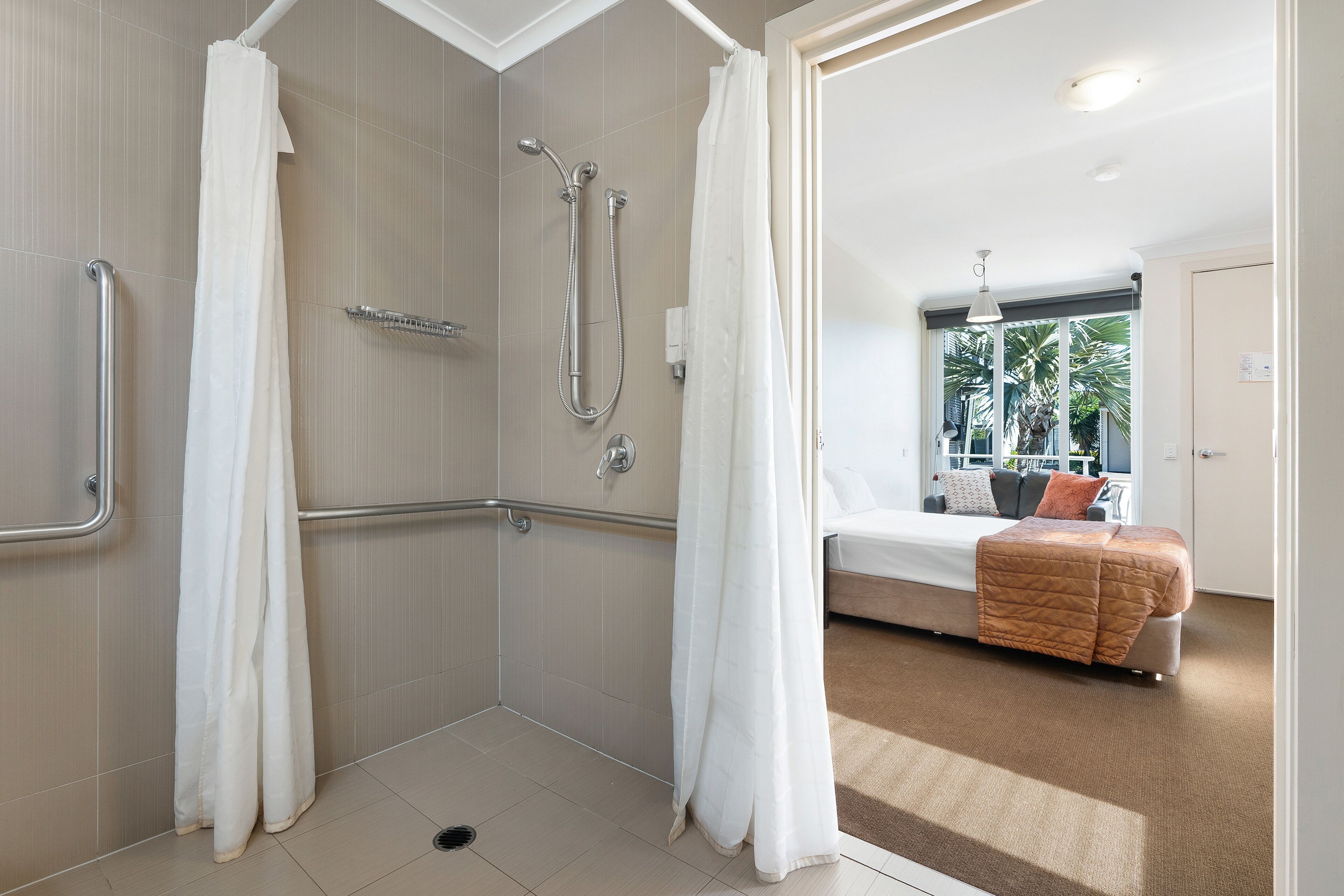 Un baño moderno con ducha, barras de apoyo y vista a un dormitorio con cama y ventana.
