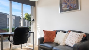 Una sala de estar moderna con un sofá de cuero negro, una silla de oficina negra y un amplio ventanal con vistas a edificios.