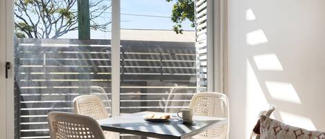 A dining area with a table and chairs, a window with blinds, and a view of a building and trees outside.
