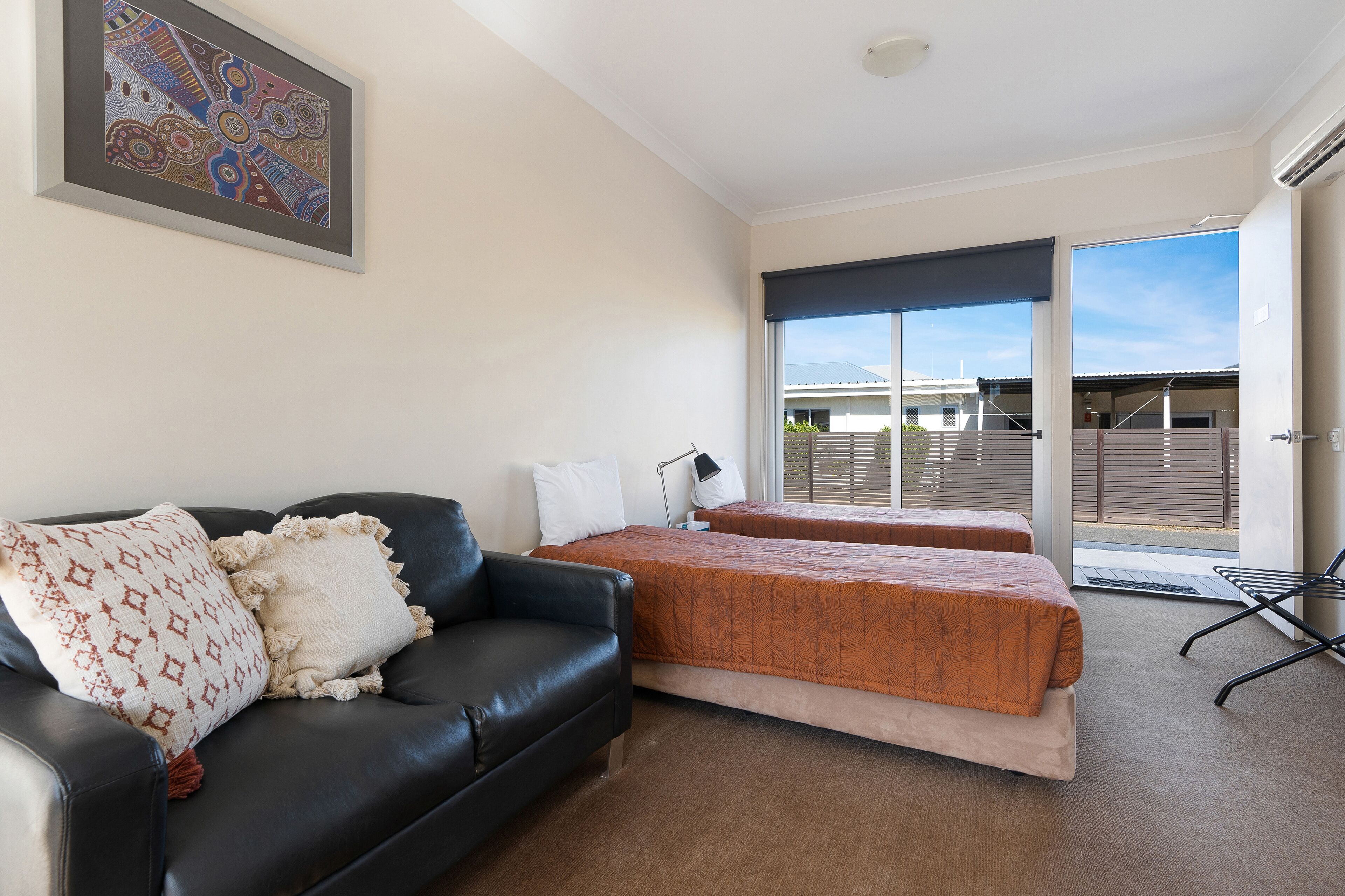 A hotel room with a black leather sofa, two beds with orange bedding, a large window with a view of the outside, and a framed artwork on the wall.