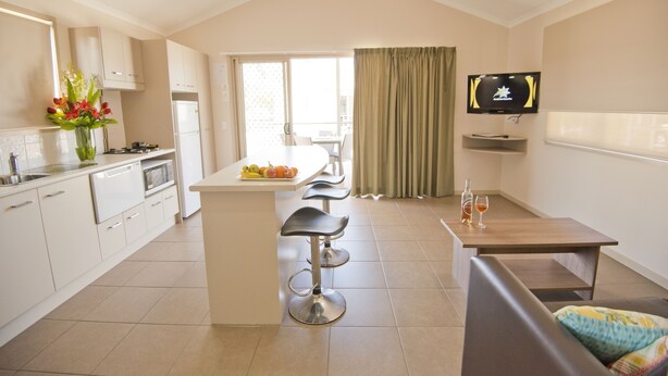 Una cocina moderna con una isla central, taburetes de bar, un comedor con mesa y sillas, un televisor montado en la pared y vistas a través de un ventanal grande.