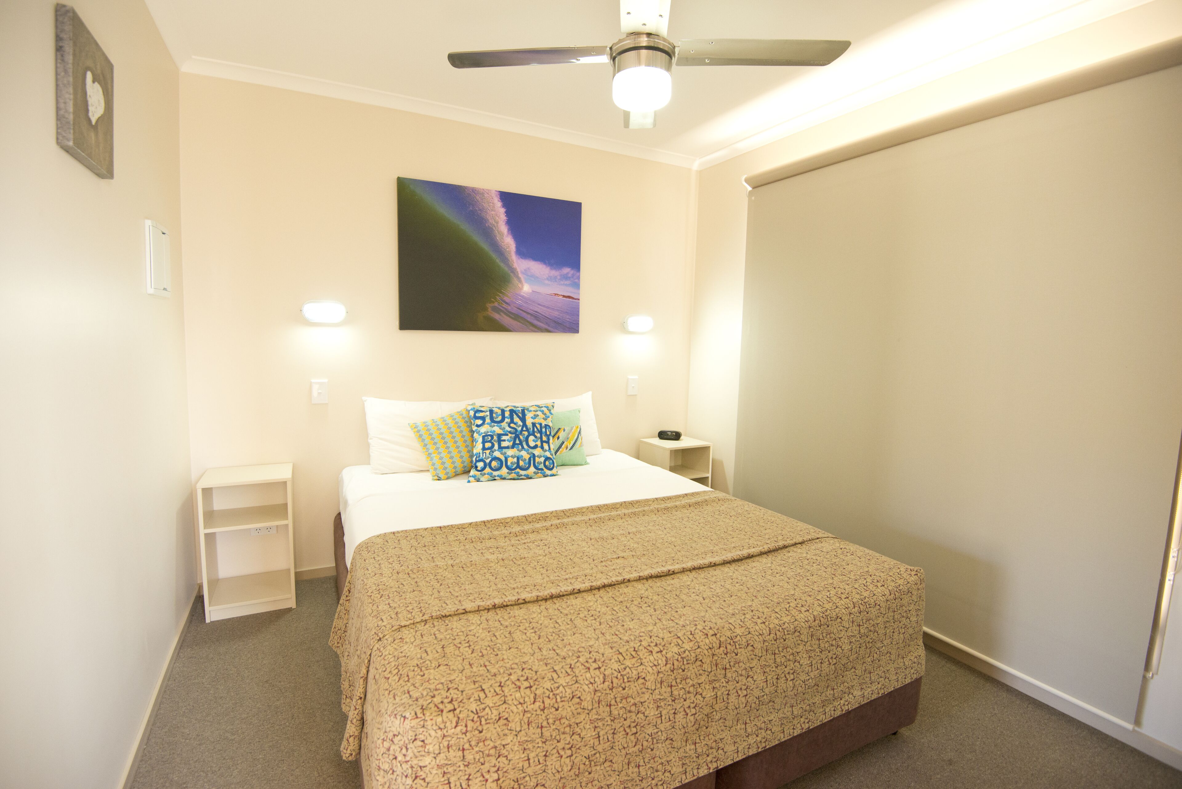 Ein Schlafzimmer mit Bett, Deckenventilator, Nachttisch, Wandleuchte und einem Strandkissen.