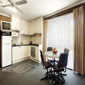 A modern kitchen with white cabinets, a microwave, a stove, and a ceiling fan. There is a dining area with a round table and four chairs, and a large window with curtains. - Paradise Holiday Apartments (Lake Bunga)