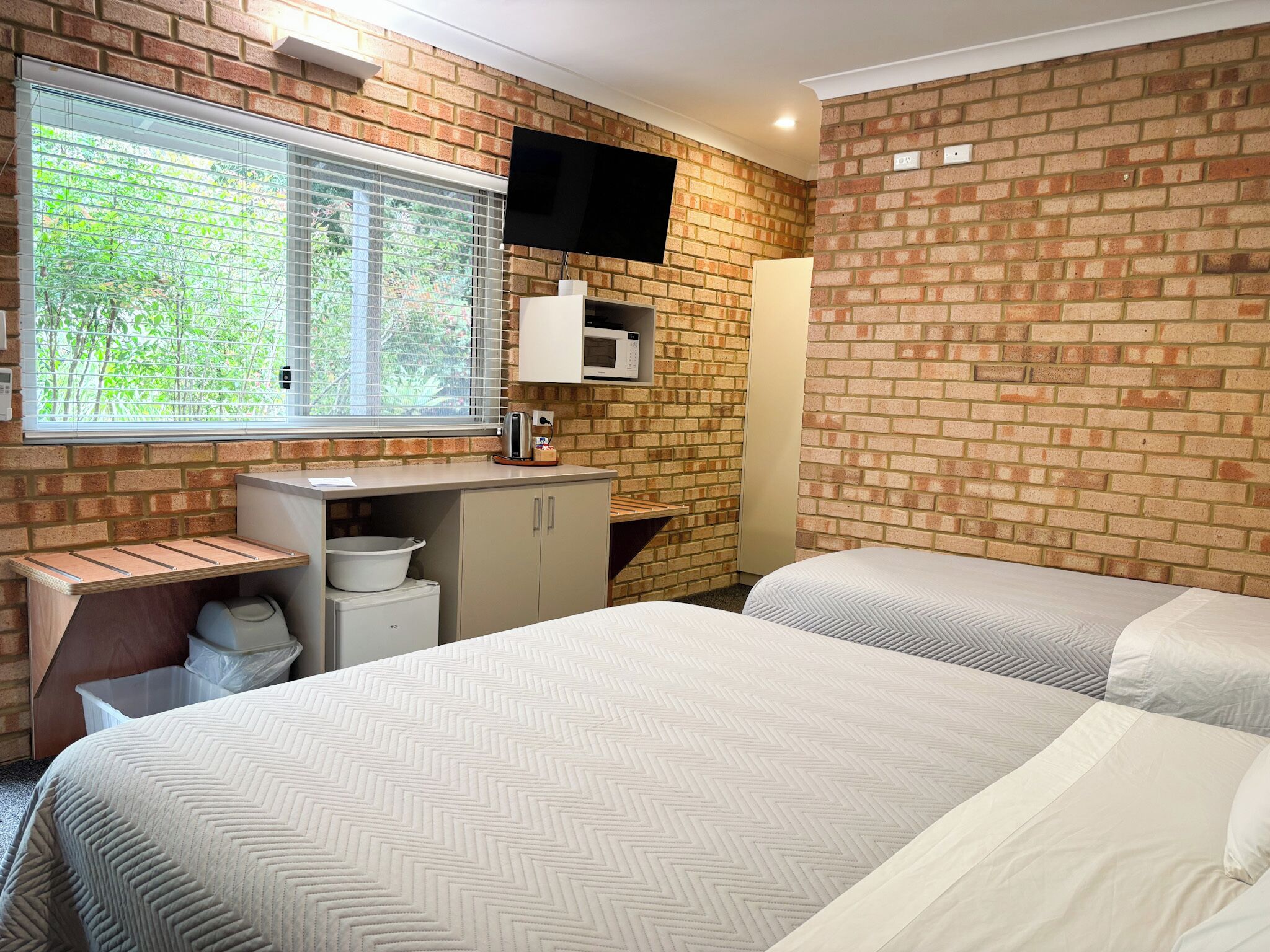 A hotel room with two beds, a TV mounted on the brick wall, a window with blinds, a sink, and a trash bin.
