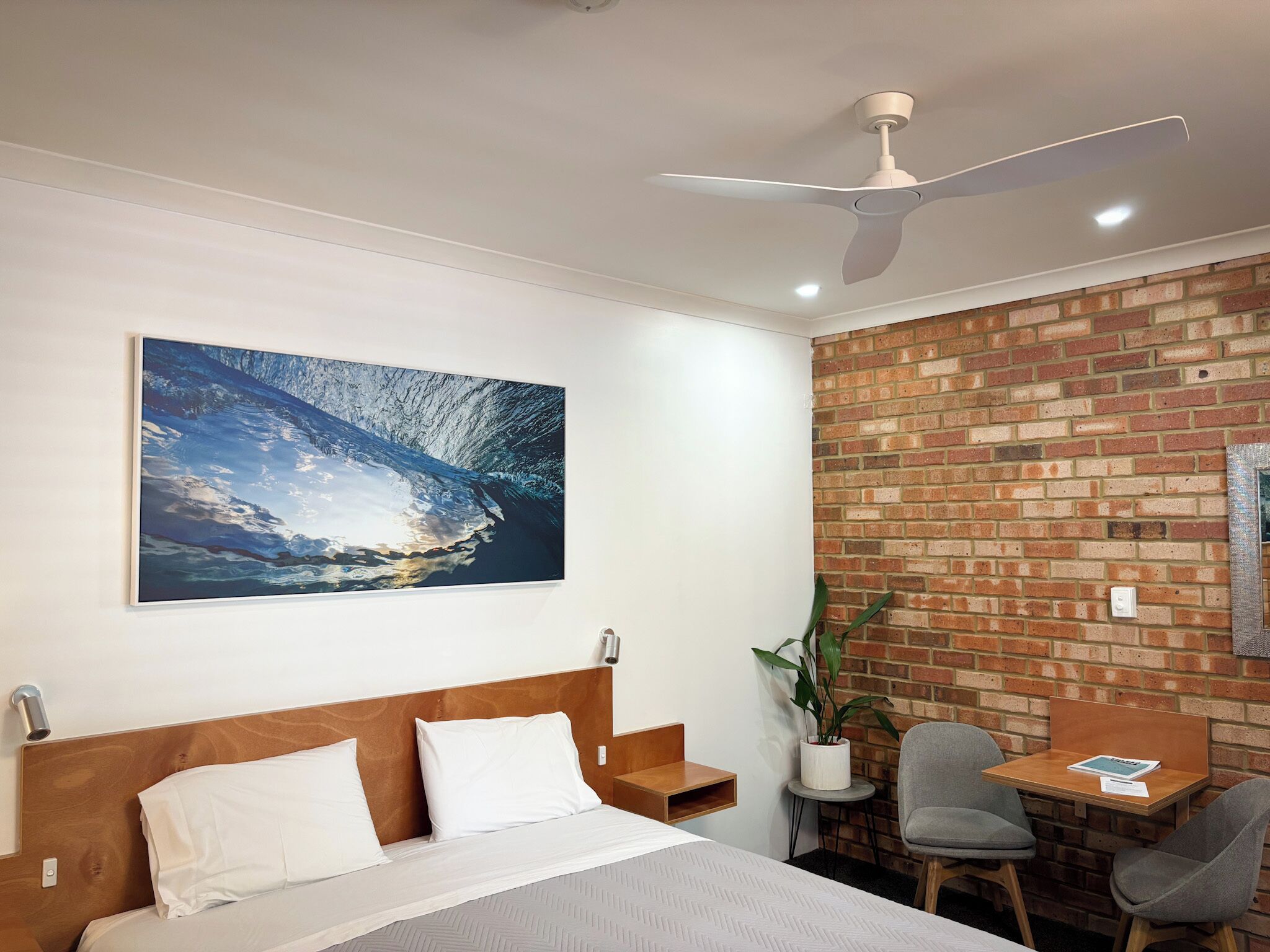A hotel room with a bed, a ceiling fan, a brick wall, a small table with a chair, and a large painting of a wave.