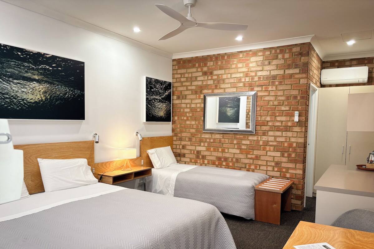 Habitación de hotel con dos camas, una pared de ladrillo decorativa, un ventilador de techo y una ventana con un marco de fotos.