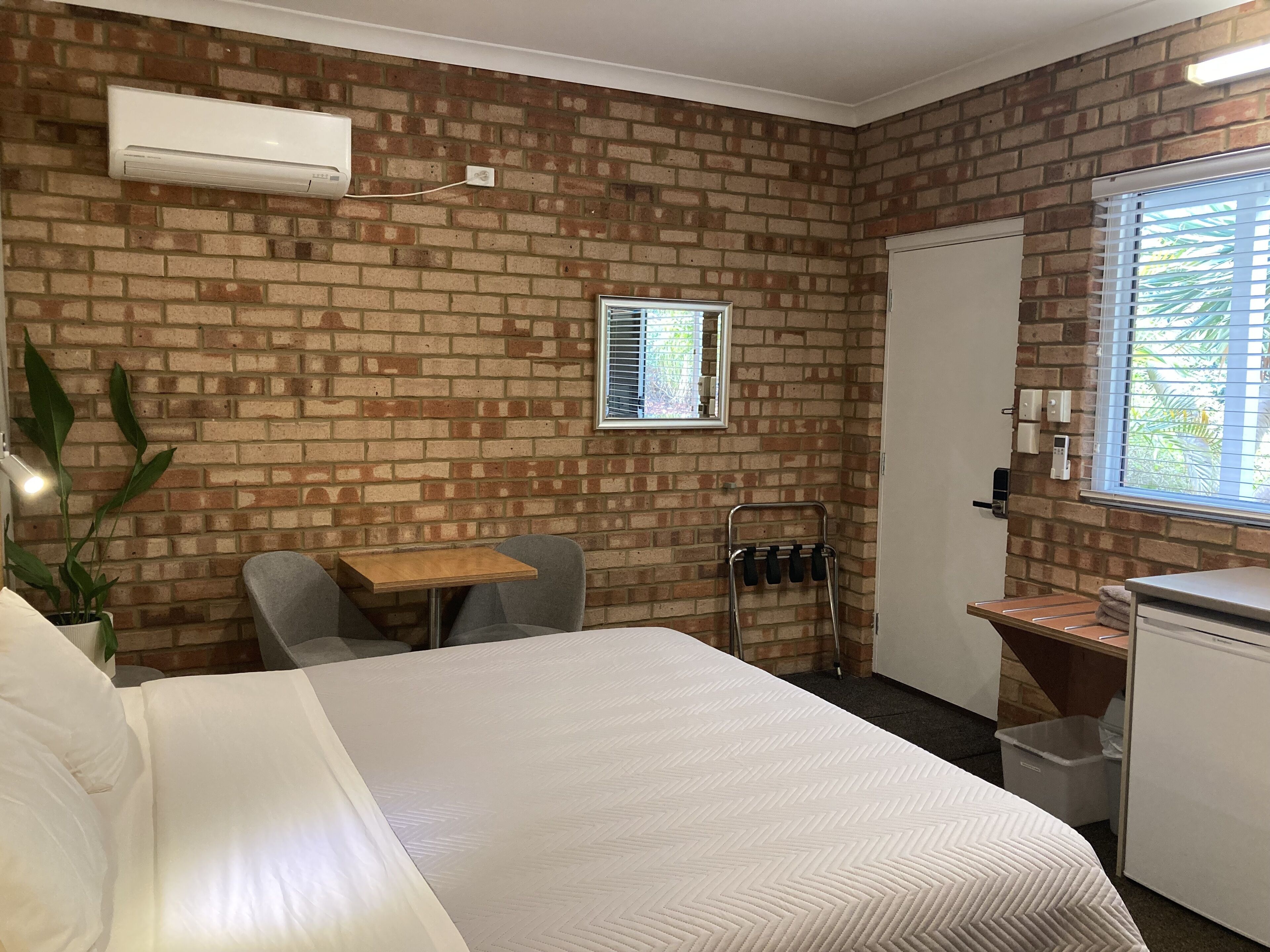 A hotel room with a brick wall, a bed, a small table, a chair, a desk, a mirror, and a window with blinds.