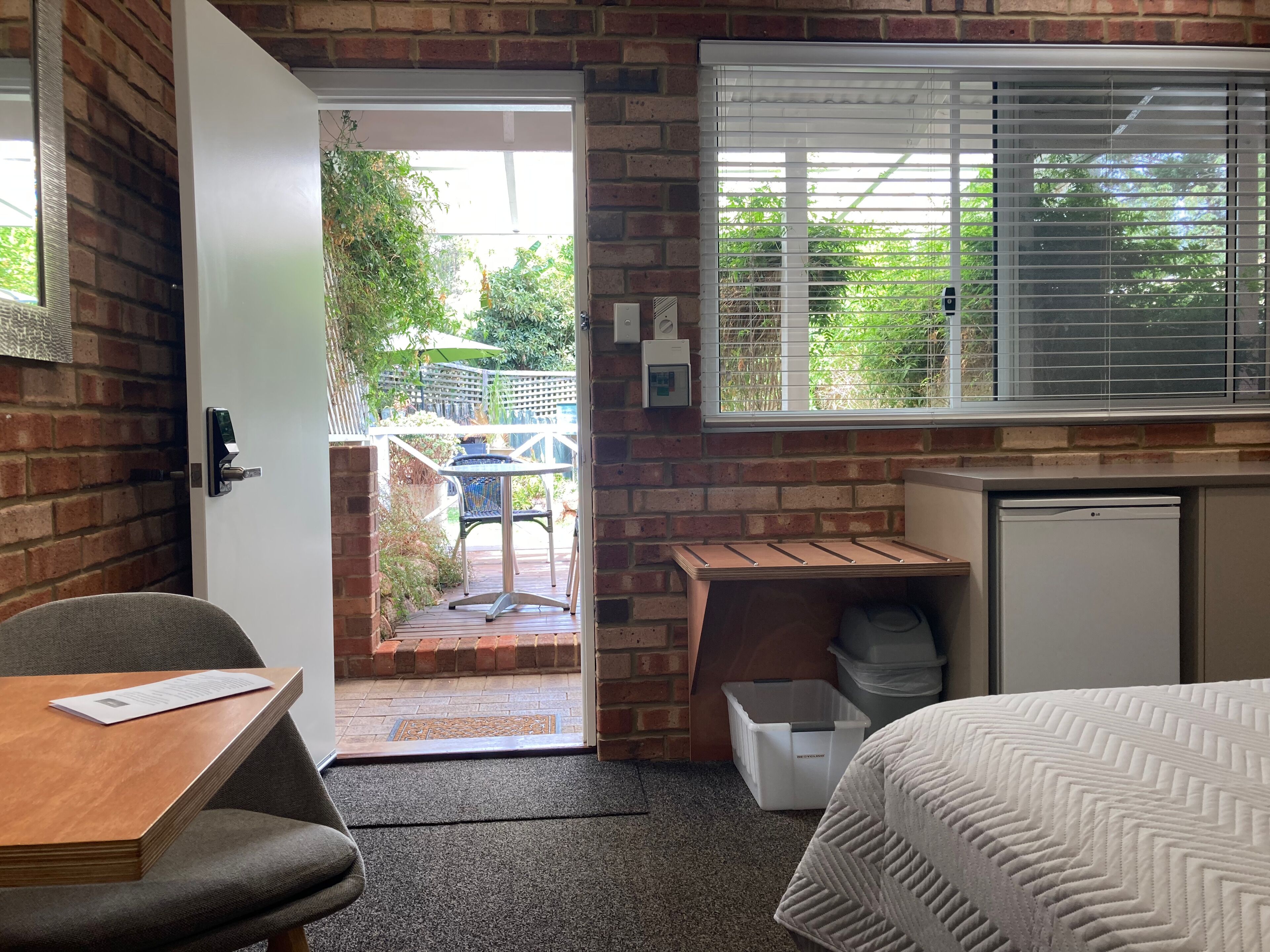 Een compacte kamer met een bakstenen muur, een klein tafeltje met een stoel, een bed, een raam met zonwering en een deur naar een buitenruimte met een tafel en stoelen.