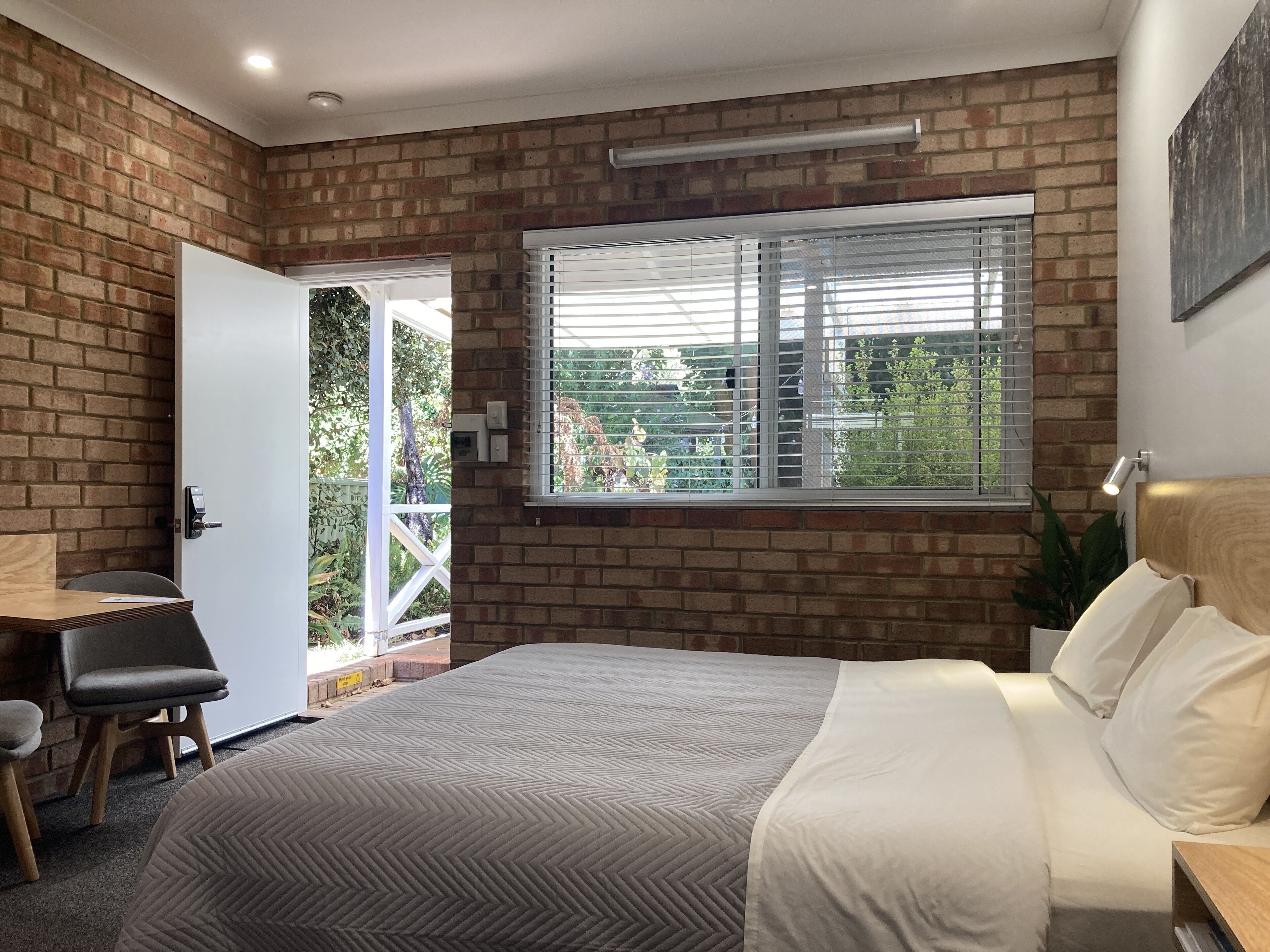 A bedroom with a brick wall, a large window with blinds, a bed with white bedding, a desk with a chair, and a small table.