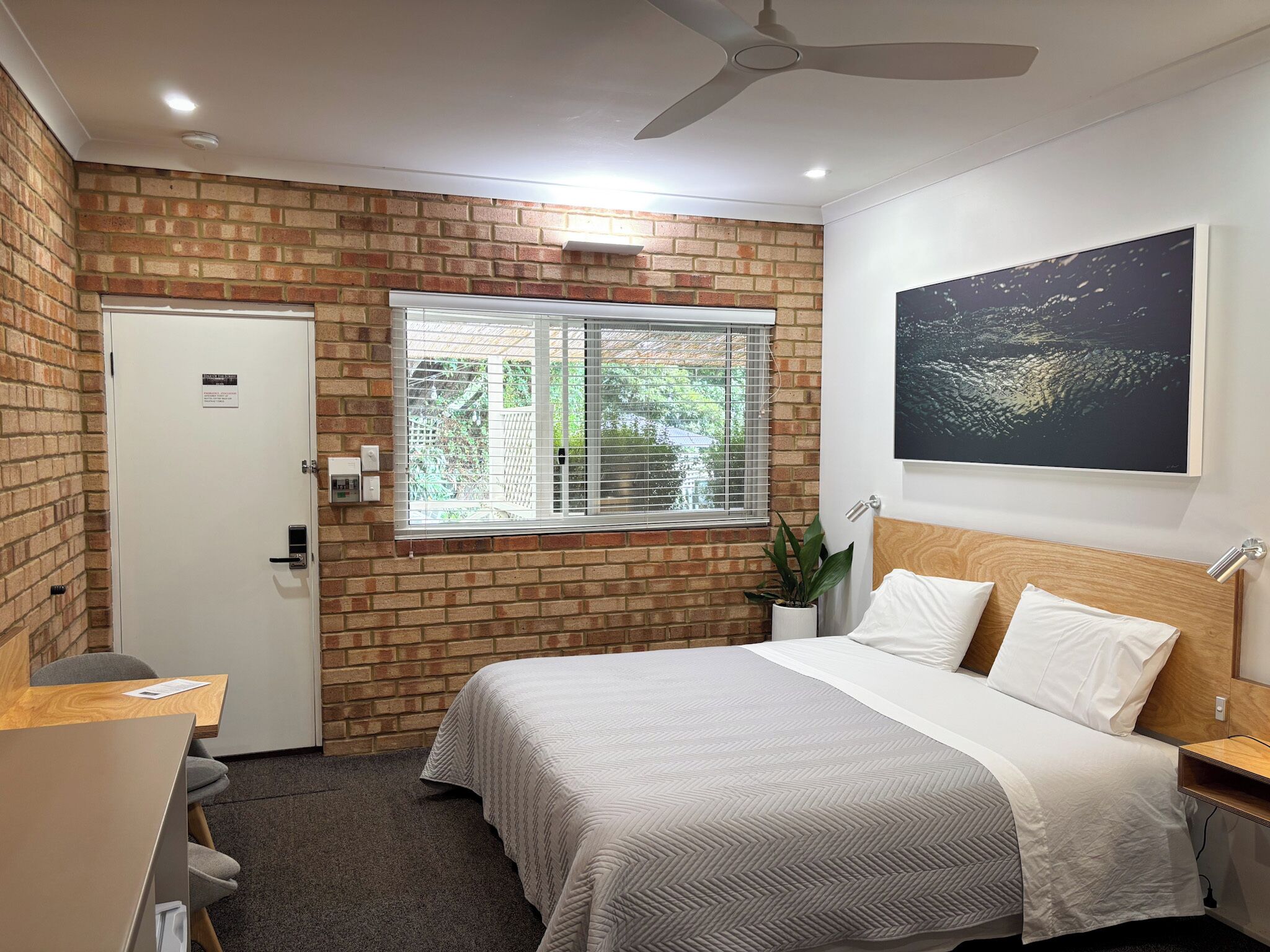 A bedroom with a brick wall, a window with blinds, a bed with white bedding, a ceiling fan, and a wall-mounted artwork.