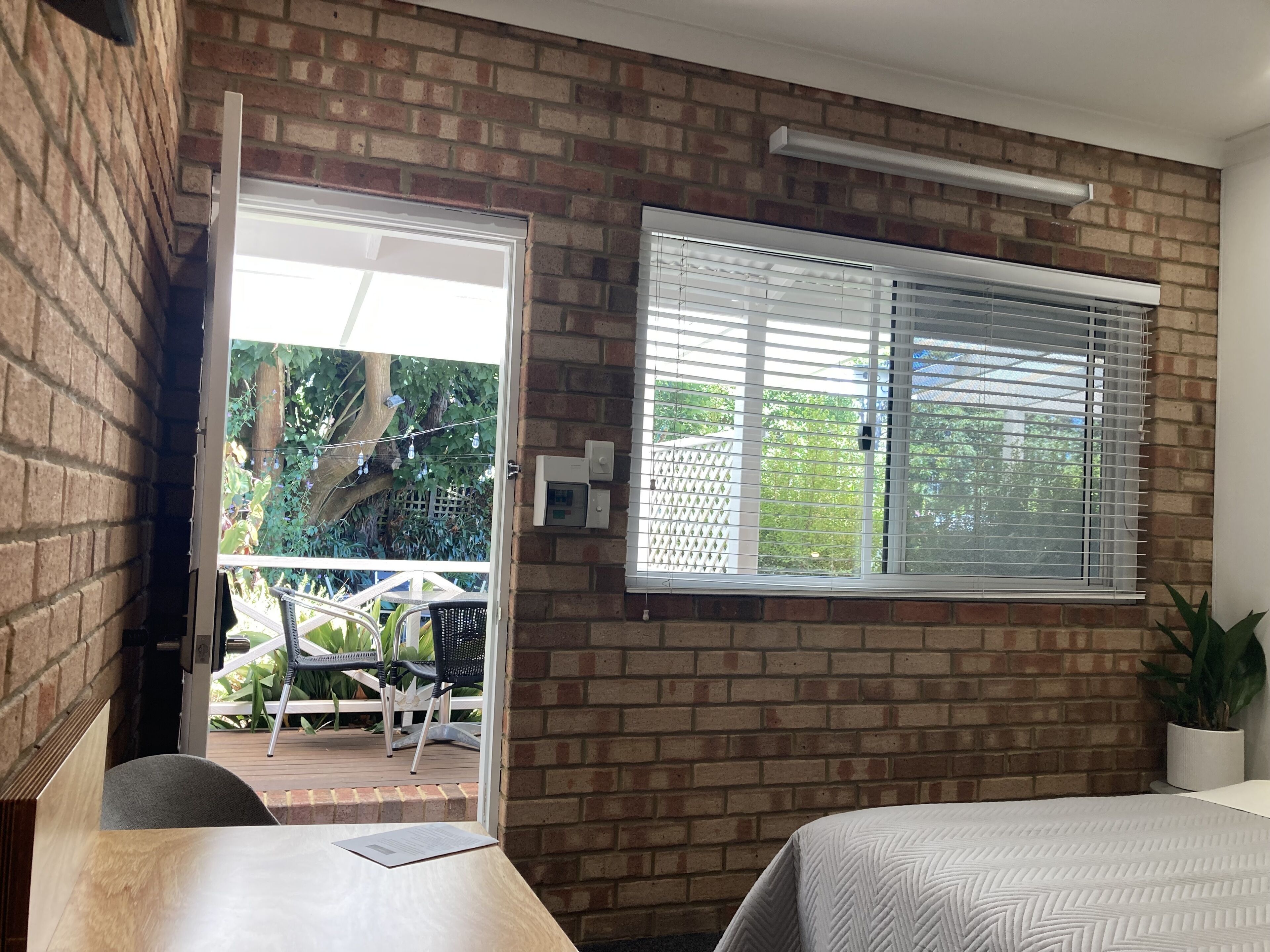 A room with a brick wall, a window with blinds, a bed, a desk with a chair, and a potted plant.