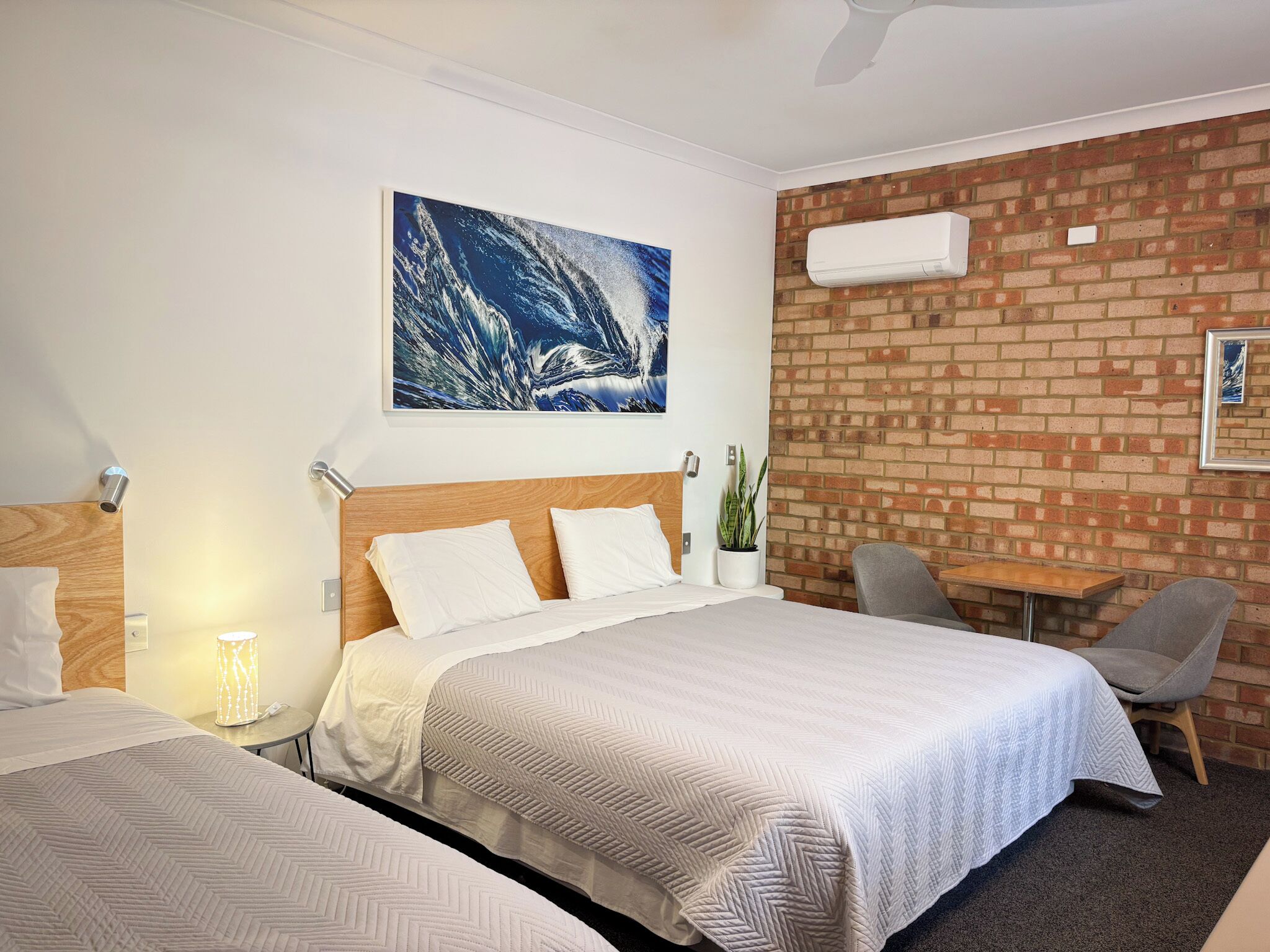 A hotel room with two beds, a brick wall, a ceiling fan, a potted plant, and a small table with a chair.