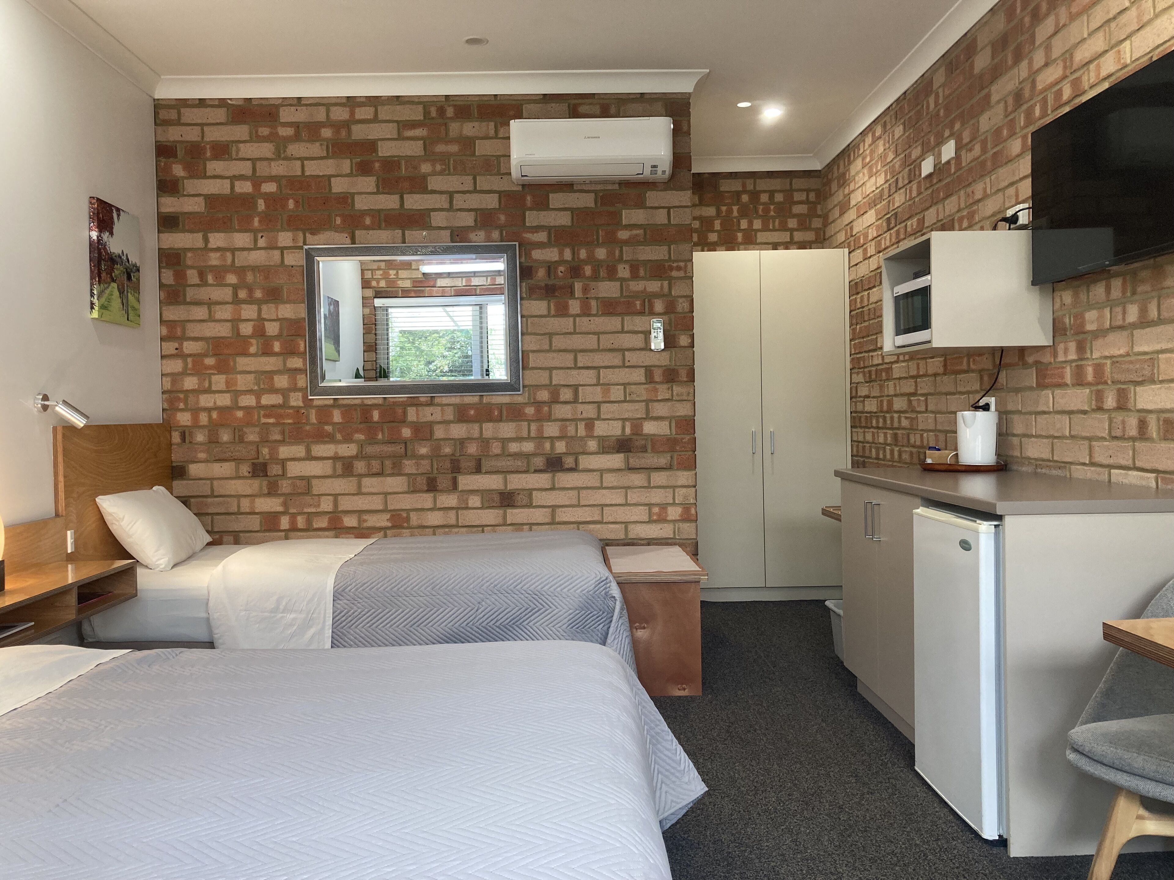 A compact hotel room with two beds, a small kitchenette, a TV, and a brick wall.