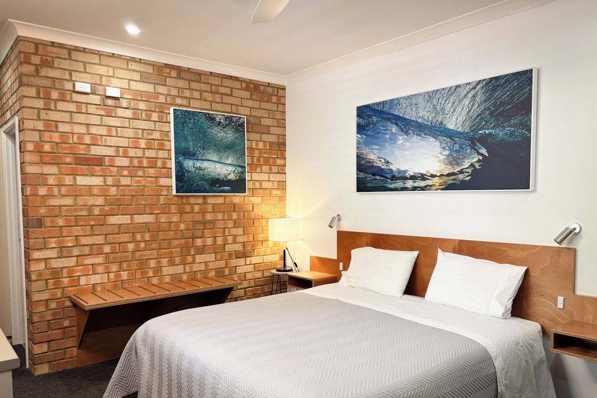Habitación de hotel con pared de ladrillo, una cama con ropa de cama blanca, una mesita de noche, una lámpara y dos cuadros enmarcados.