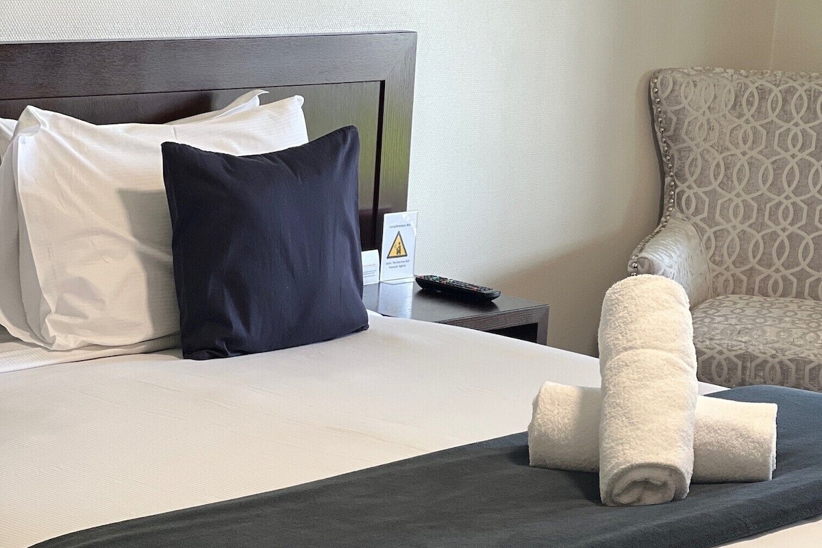 A neatly made bed with white and dark grey linens, a remote control on the nightstand, and a patterned armchair in the background.