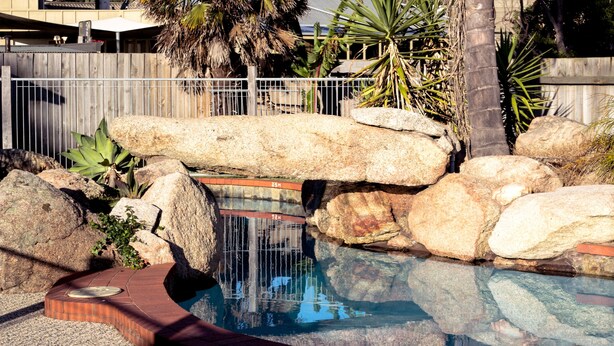Zona de piscina con rocas grandes, un muelle de madera y una valla.