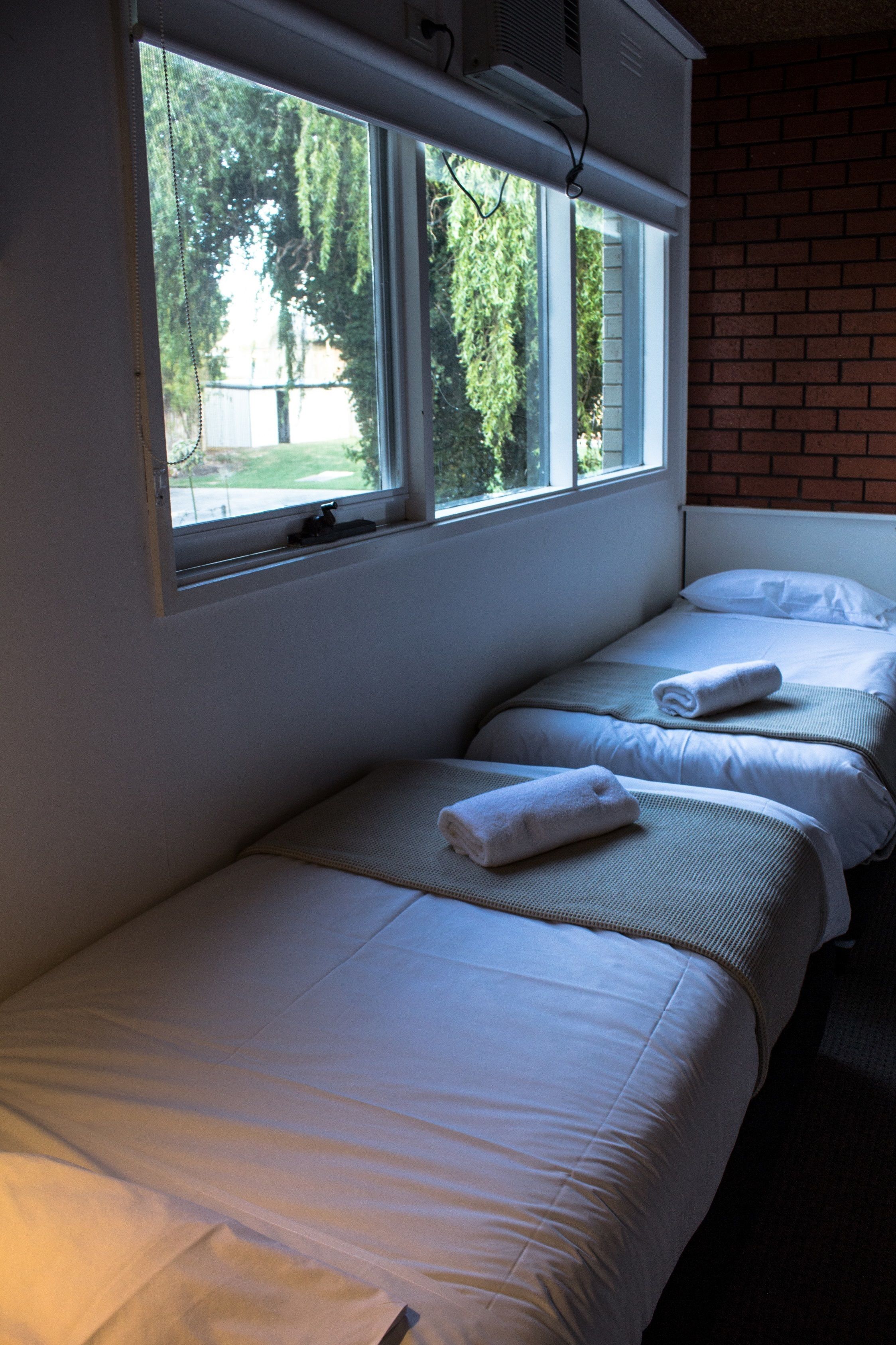 A room with two single beds, each with a white mattress and pillow, and a window providing a view of trees outside.