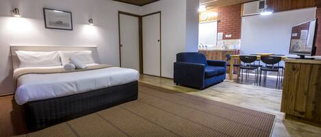 A hotel room with a bed, a blue armchair, a small dining table with chairs, a kitchenette, and a whiteboard.