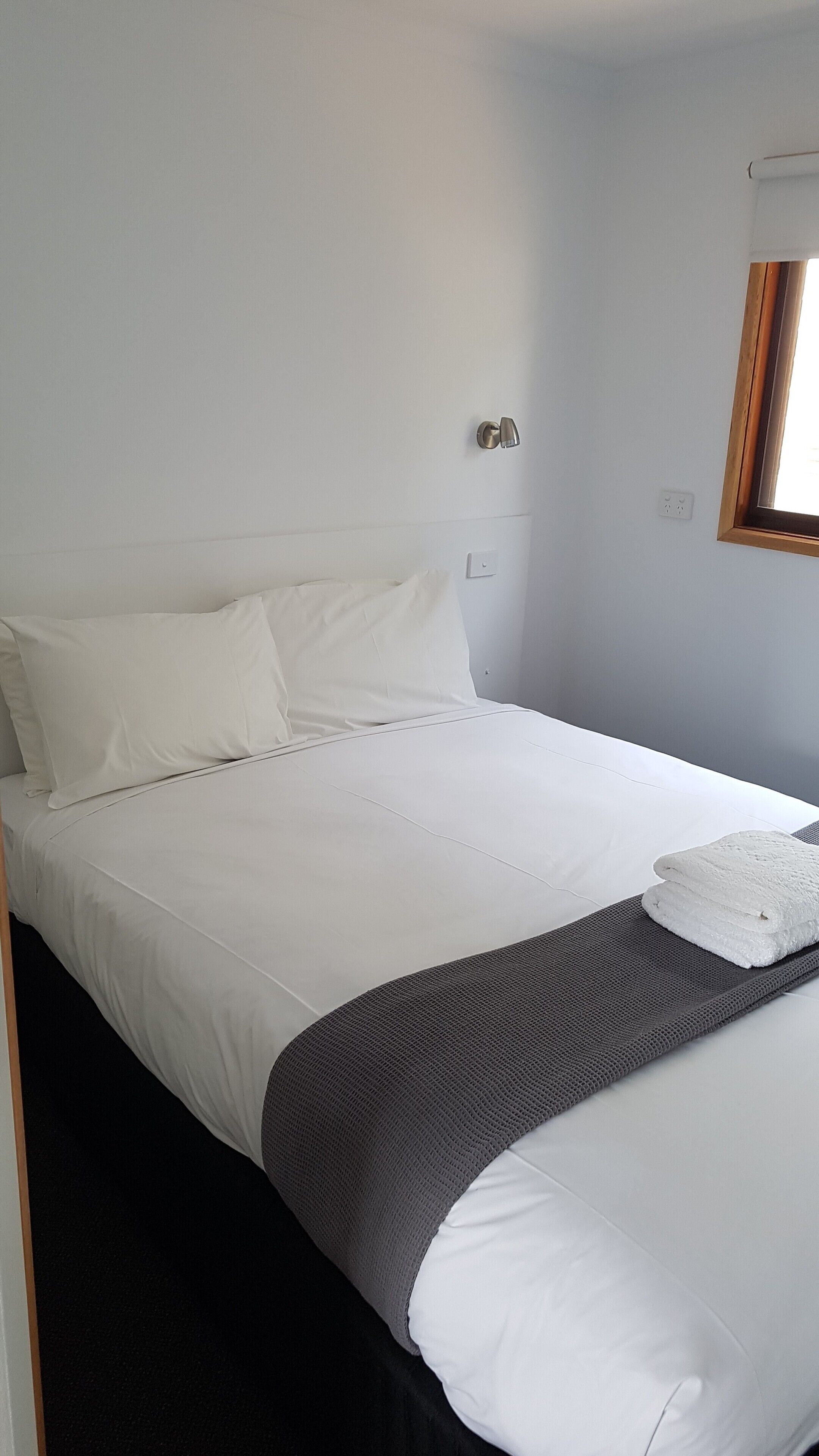 A neatly made bed with white linens and a gray blanket, a wall-mounted light fixture, and a window with a wooden frame.