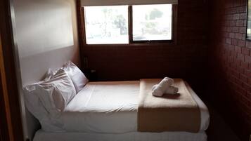 A single bed with white linens, a window with a view of the outside, and a brick wall.
