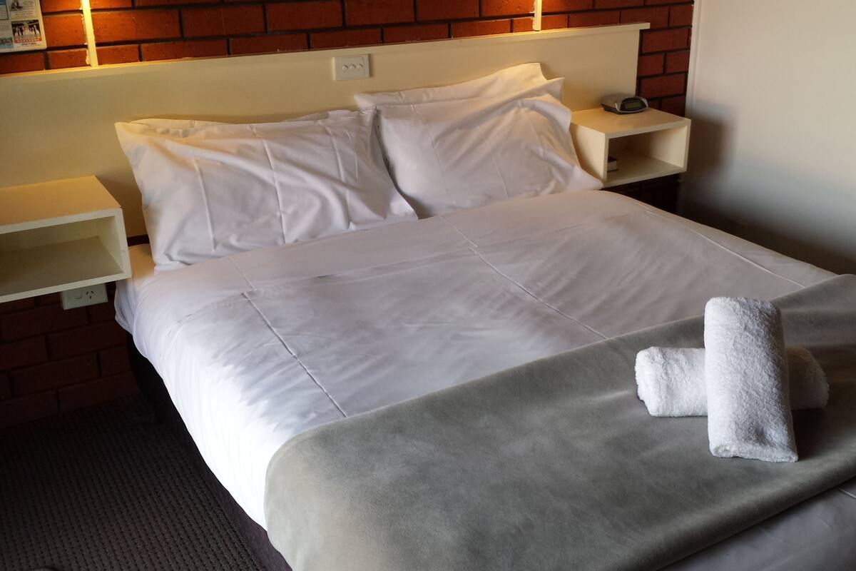A neatly made bed with white linens and a grey blanket, a white towel on the bed, and a brick wall in the background.