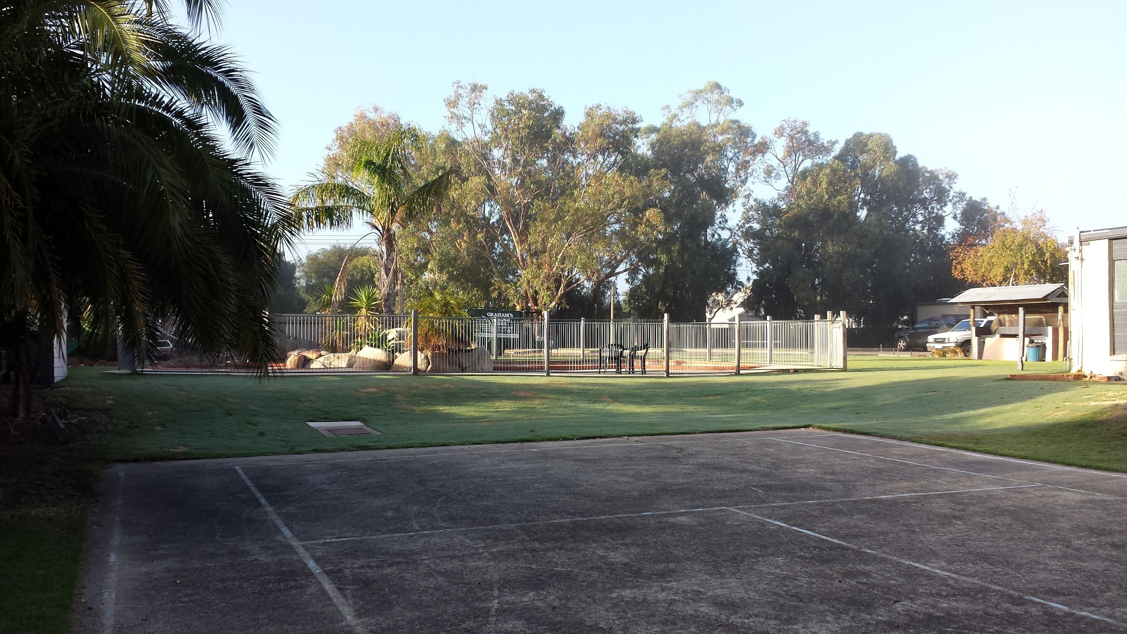 A fenced outdoor area with a paved section, surrounded by trees and a building.