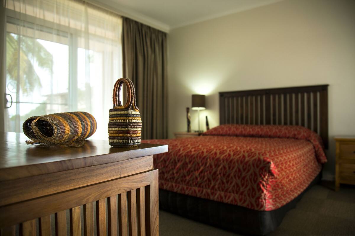 A hotel room with a wooden desk, a patterned red bedspread, a lamp, and a window with curtains.
