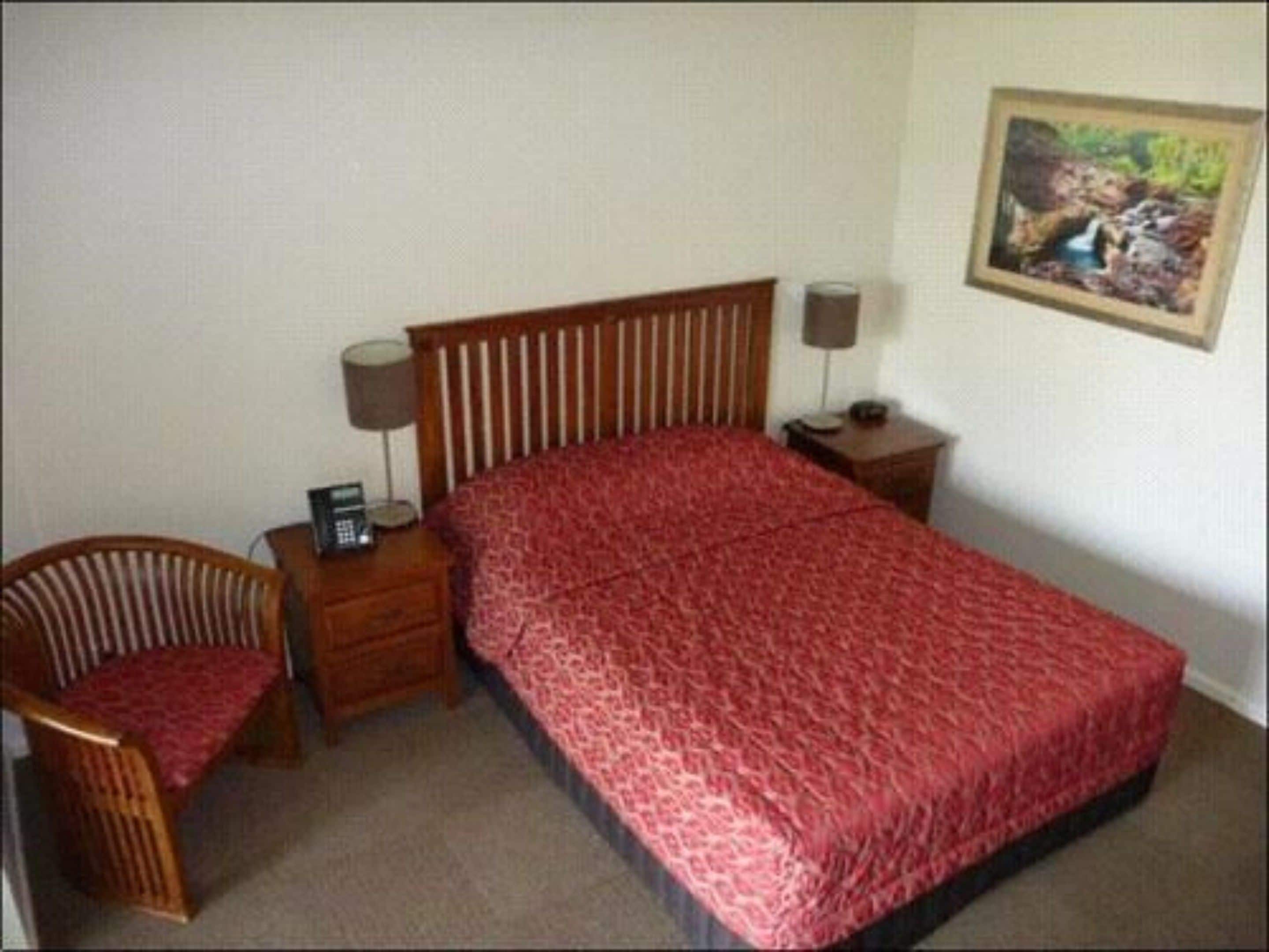 Een slaapkamer met een bed, een stoel, een nachtkastje met een lamp en een schilderij aan de muur.