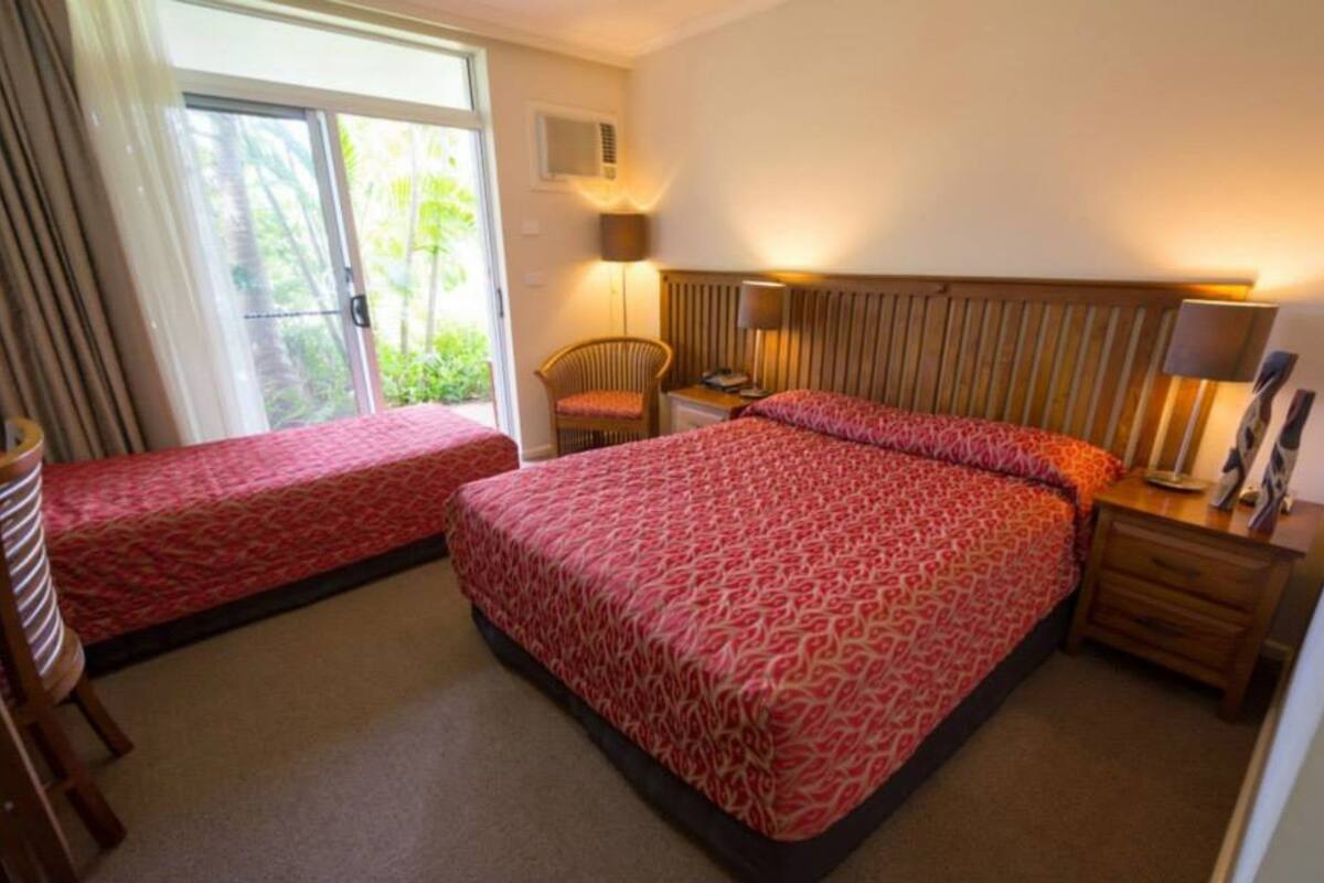 A hotel room with two beds, a chair, a nightstand, a lamp, and a sliding glass door to the outside.