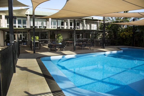 A swimming pool with a sunshade structure, outdoor seating, and a building with multiple floors in the background. - Walkabout Lodge (Nhulunbuy)
