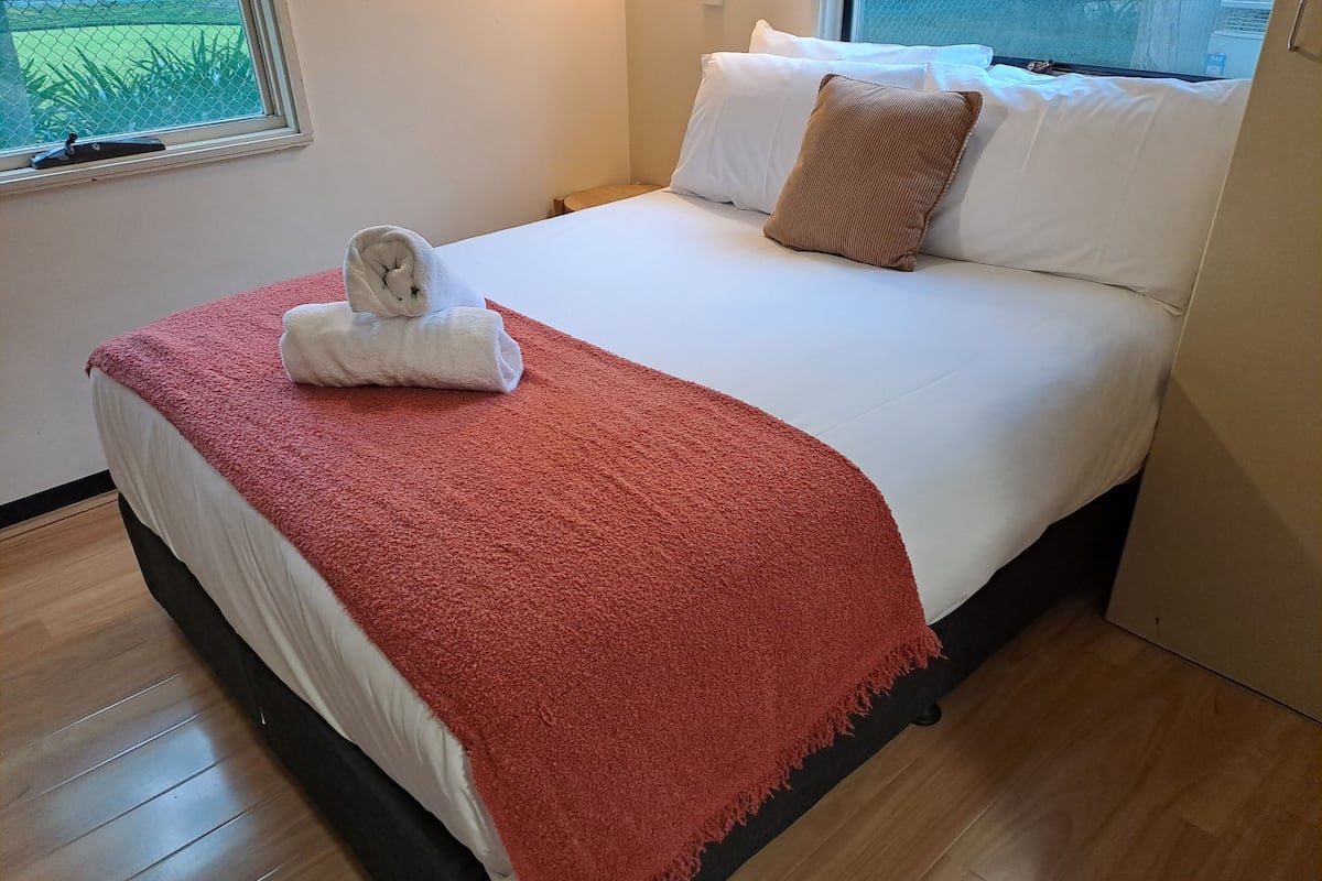 Un lit bien fait, avec une couette blanche, un plaid rouge et une serviette enroulée posée sur le pied de lit.